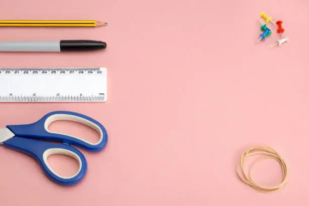 Office or school supplies including a pair of scissors, ruler, marker pen, a pencil, thumbtacks, and rubber bands on pink background with copy space
