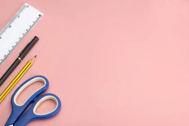 Scissors, a pencil, a ballpoint pen, and a plastic ruler on pink backdrop with copy space