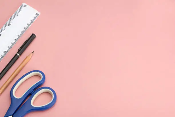 Blue-handled scissors, a pencil, a ballpoint pen, and a plastic ruler on pink background with copy space