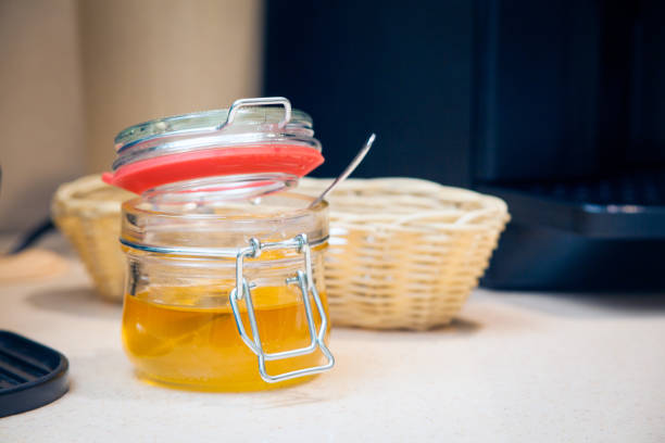 A close-up of natural golden honey in a glass transparent jar with a spoon is on the breakfast table, in the background a wicker basket