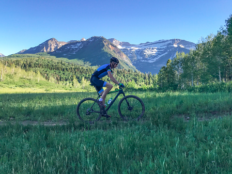 A man rides his mountain bike through a meadow with a high alpine peak behind.