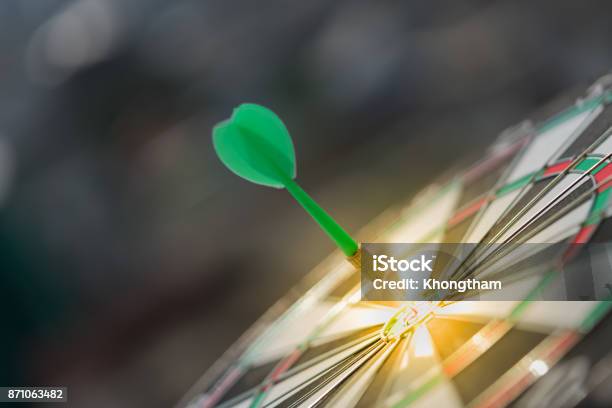 Green Dart Arrow Hitting In The Target Center Of Dartboard With Modern City And Sunset Background Stock Photo - Download Image Now