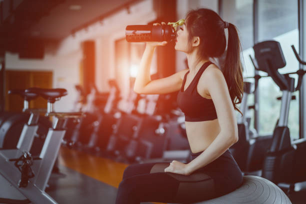 giovane donna asiatica che beve proteine scuotono dalla bottiglia dopo l'allenamento per i muscoli nella palestra fitness sano stile di vita bodybuilding seduto su palla yoga - whey protein concentrate foto e immagini stock
