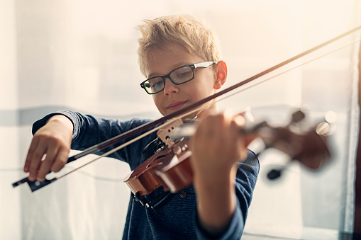 Closeup portrait of a focused little boy playing violin. The boy is aged 7 and is playing in a sunny room.\n