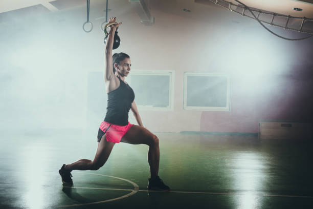 donna atletica che fa affondo con la campana del bollitore in palestra. - kettle bell sport women muscular build foto e immagini stock