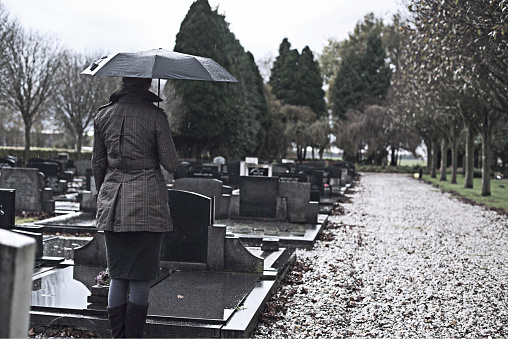 Widow woman visiting grave of lost relative, husband or child. in the rain under umbrella. Dark setting