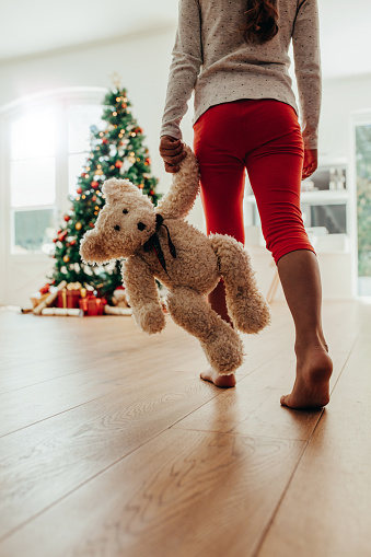 Low section of young girl holding teddy bear walking towards Christmas tree. Decorated Christmas tree and gifts arranged in living room.