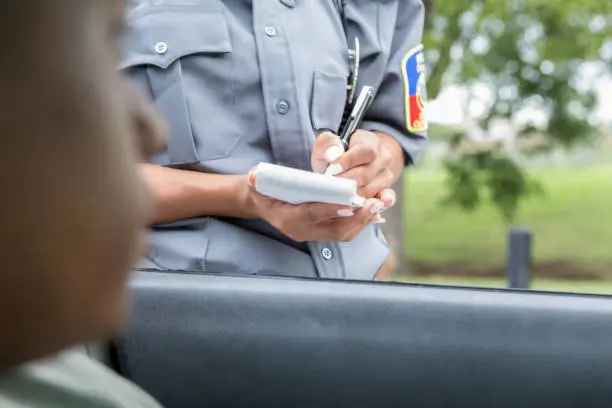 In this closeup from inside the vehicle, an unrecognizable driver waits for an unrecognizable police officer as she writes him a speeding ticket.  The officer can be seen holding a pad and pen and writing the ticket.