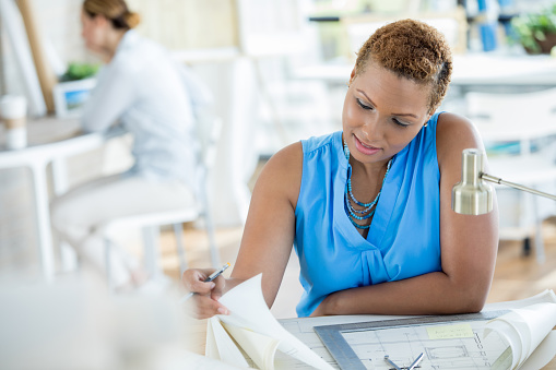 Mid adult African American architect concentrates while reviewing a blueprint. A colleague is working in the background.