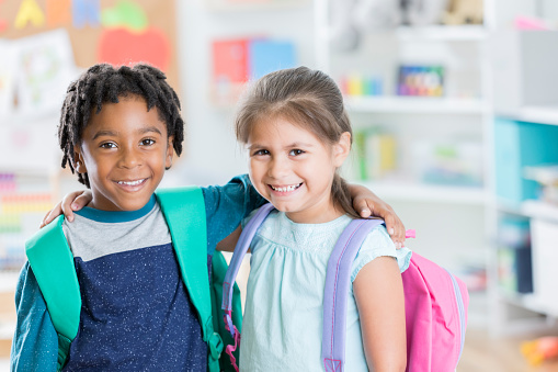 Two elementary students, a boy and a girl, stand in their school and smile for the camera.  They are wearing backpacks as they each put an arm on the other's shoulder in friendship.