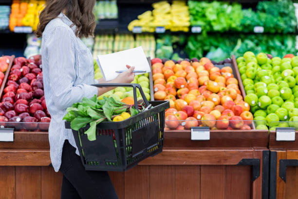 Unrecognizable woman shops for produce in supermarket Young woman carries a shopping basket filled with fresh produce. She is shopping for fresh fruit and vegetables in a grocery store. freshness stock pictures, royalty-free photos & images