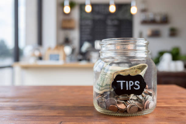""Tips"" jar on coffee shop table Foreground focus is on a glass jar labeled "tips" in chalk.  The jar is sitting to one side on a rustic wooden table, full of coins and bills, with coffee shop scenery in the background. begging social issue photos stock pictures, royalty-free photos & images