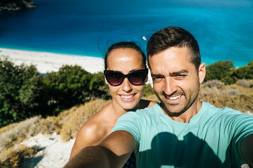 Beautiful, young couple taking selfie with blue sea and pebbly beach in background.