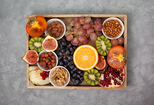 Stock photo showing close-up, elevated view of wooden charcuterie board covered with prepared sliced and chopped ingredients including crackers, ham, prosciutto and salami roses, cucumber, lime half, cherry tomatoes, Brie, kiwi, Cheddar, red grapes, ramekin of honey and honey dipper.