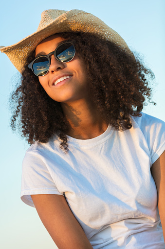 Beautiful happy mixed race African American female girl teenager young woman wearing reflective aviator sunglasses and cowboy hat outside at golden sunset or sunrise