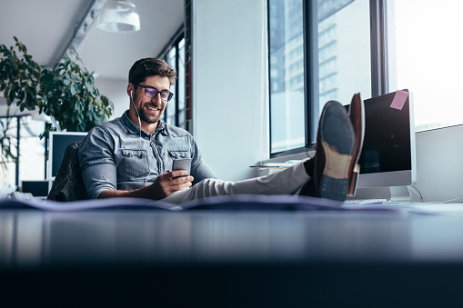 Businessman using mobile phone in the office with feet on the table. Male designer listening music in break time.