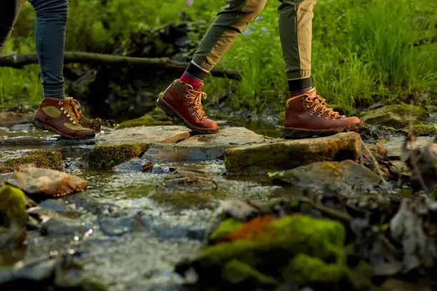 Woman hiker crossing a small forest stream in a close up low angle view of her feet in leather boots on natural rock stepping stones
