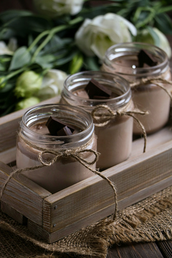 tasty and sweet dessert - chocolate panna cotta in three glass jars close up,  tied with rope, slices of dark chocolate, on a vintage wooden tray, white eustoma flowers at the back side