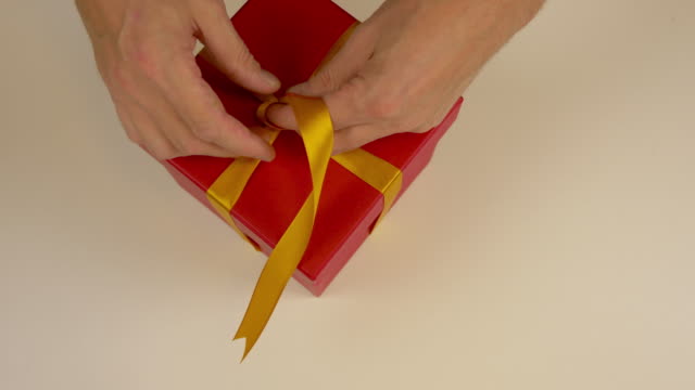 Close up top view high angle. Mens hands straighten a gold bow from a satin ribbon on a gift box. Preparing for the gift giving event. Gift box with yellow bow. Red carton box.
