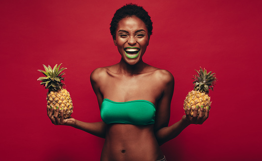 Laughing young african woman with pineapples in hand. Female fashion model with pineapples laughing over red background.