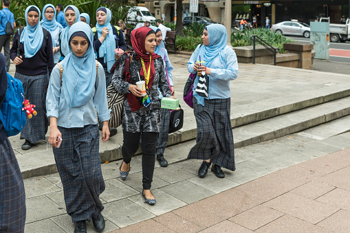 Sydney, Australia - March 23, 2017: Group of young female Muslims walking in town. Covered with azur-colored head-and-hair covering scarves and long dark skirts as a uniform. Female teacher with maroon scarf.