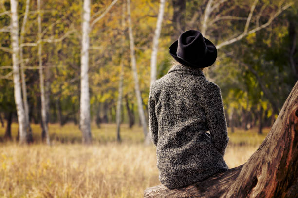 istock Girl in a hat sitting on a log in the autumn forest. Back view. 871308630
