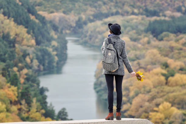 istock Girl with a backpack and a hat standing on a hill. River and forest below. Back view 871308662