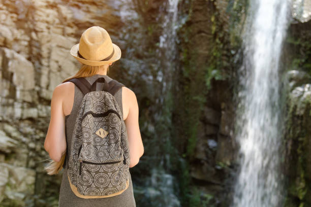 istock Girl with backpack and in a hat looking at a waterfall. View from the back 871312020