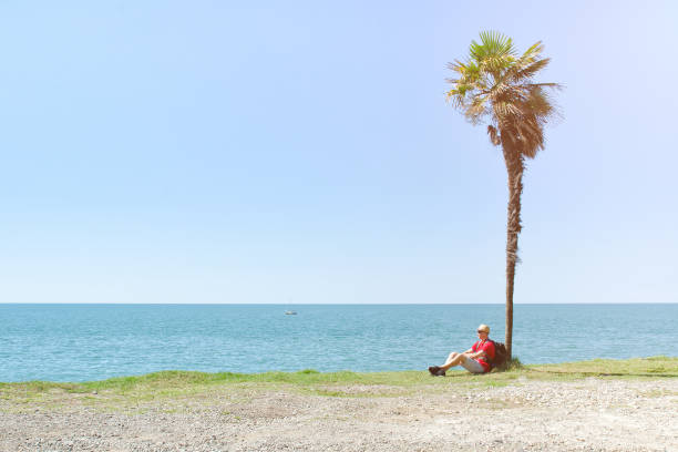 istock Man with backpack sits under a tall palm tree on background of sea and blue sky 871320102