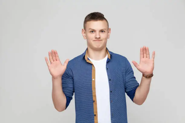 Photo of Hands up. Young boy wearing white tshirt and blue shirt looking at camera and give up