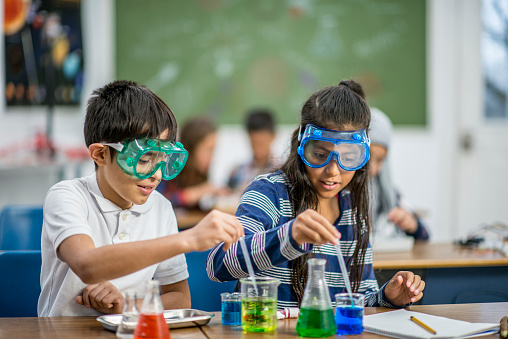 A multi-ethnic group of elementary school students are indoors in a classroom. They are wearing casual clothing. An Ethnic boy and girl are wearing protective goggles, and doing experiments with colored liquids.