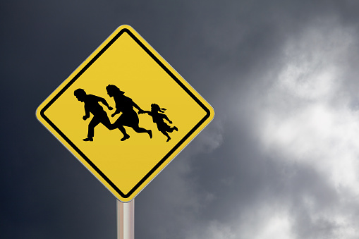 Diamond-shaped crossing sign with yellow background and black border with a family running to let drivers know that illegal migrants may be crossing before their wheels.