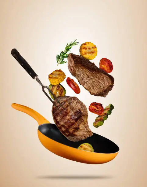 Photo of Flying beef steaks with grilled vegetable on pan, separated on beige background