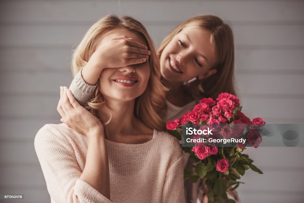 Mom and daughter Pretty teenage daughter is holding flowers and covering her mom's eyes while making a surprise, both are smiling Mother Stock Photo