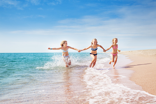Portrait of three preteen girls, happy friends, holding hands, running in sand and waves of sunny beach making splashes