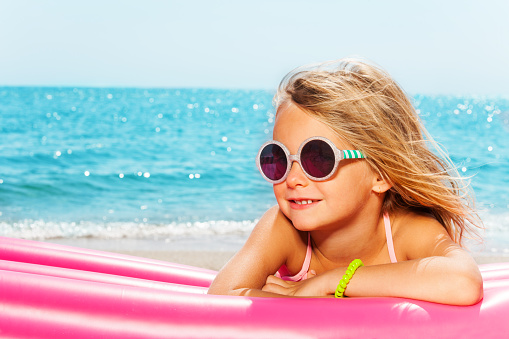 Portrait of little blond girl in sunglasses, sunbathing on pink inflatable lounge against blue sea