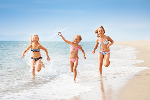 Three age-diverse girls, happy friends in swimsuits, running on the seashore with airplane model in summer
