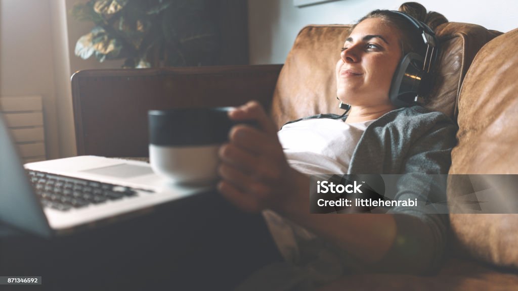 Woman listening to music in headphones on cozy sofa Pretty smiling girl listening to music from headphones with pleasure at coworking office, lying on sofa, using laptop, drinking hot coffe from nice cup and resting. Headphones Stock Photo