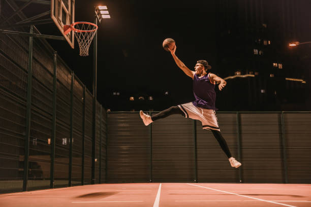Full length portrait of stylish young basketball player in cap jumping and shooting a ball through the hoop while playing outdoors at night