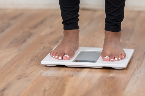 A pair of female feet with painted toes can be seen standing on a white bathroom scale.  The scale sits on a wooden floor and there is copy space.