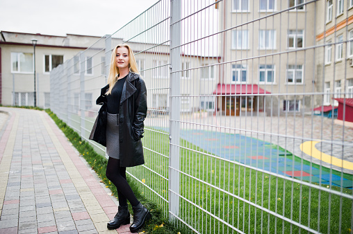 Blonde fashionable girl in long black leather coat against iron fence at street.