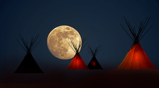 First Nation teepee camp under full moons howing a dim light inside some of the teepees.