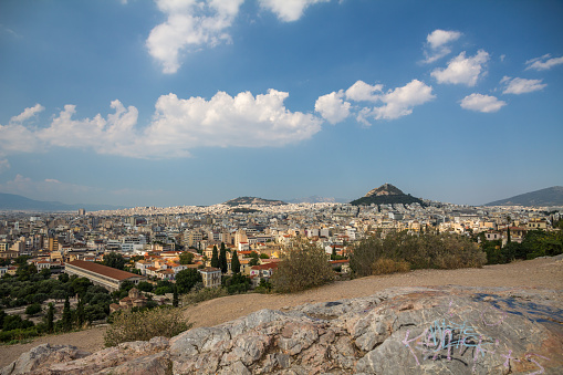 The City of Athens spreads out below Mars Hill where rocks in the foreground have been adorned by grafitti.  Light white clouds float in the azure blue sky.