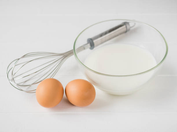 istock Two eggs, a whisk and a bowl of milk on white wooden table. 871454532