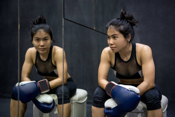 Young Asian woman boxer sitting near the mirror and posing with blue boxing gloves Young Asian woman boxer sitting near the mirror and posing with blue boxing gloves boxercise stock pictures, royalty-free photos & images