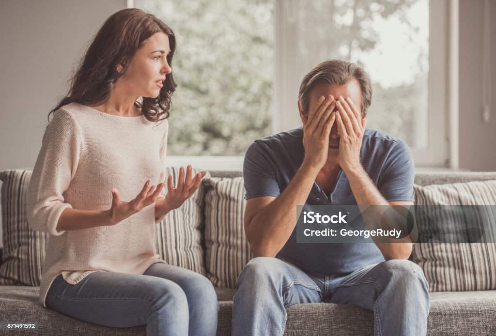 Couple having a quarrel Couple of adults are quarreling while sitting on couch at home, man is covering his face Couple - Relationship Stock Photo