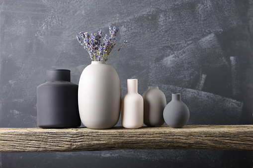 Home decor - various neutral colored vases with lavander bouquet on rough distressed wooden shelf against grey wall.