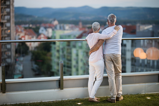 Rear view of senior couple standing embraced on a penthouse terrace and looking at view.