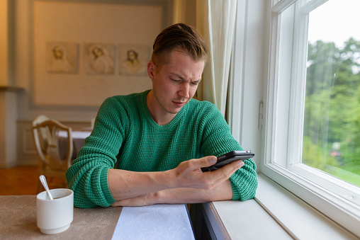 Portrait of young handsome man with blond hair wearing green sweater and relaxing by the window at home in Turku Finland