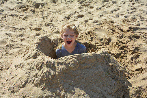Boy has dug himself in in sand and smiles forwards